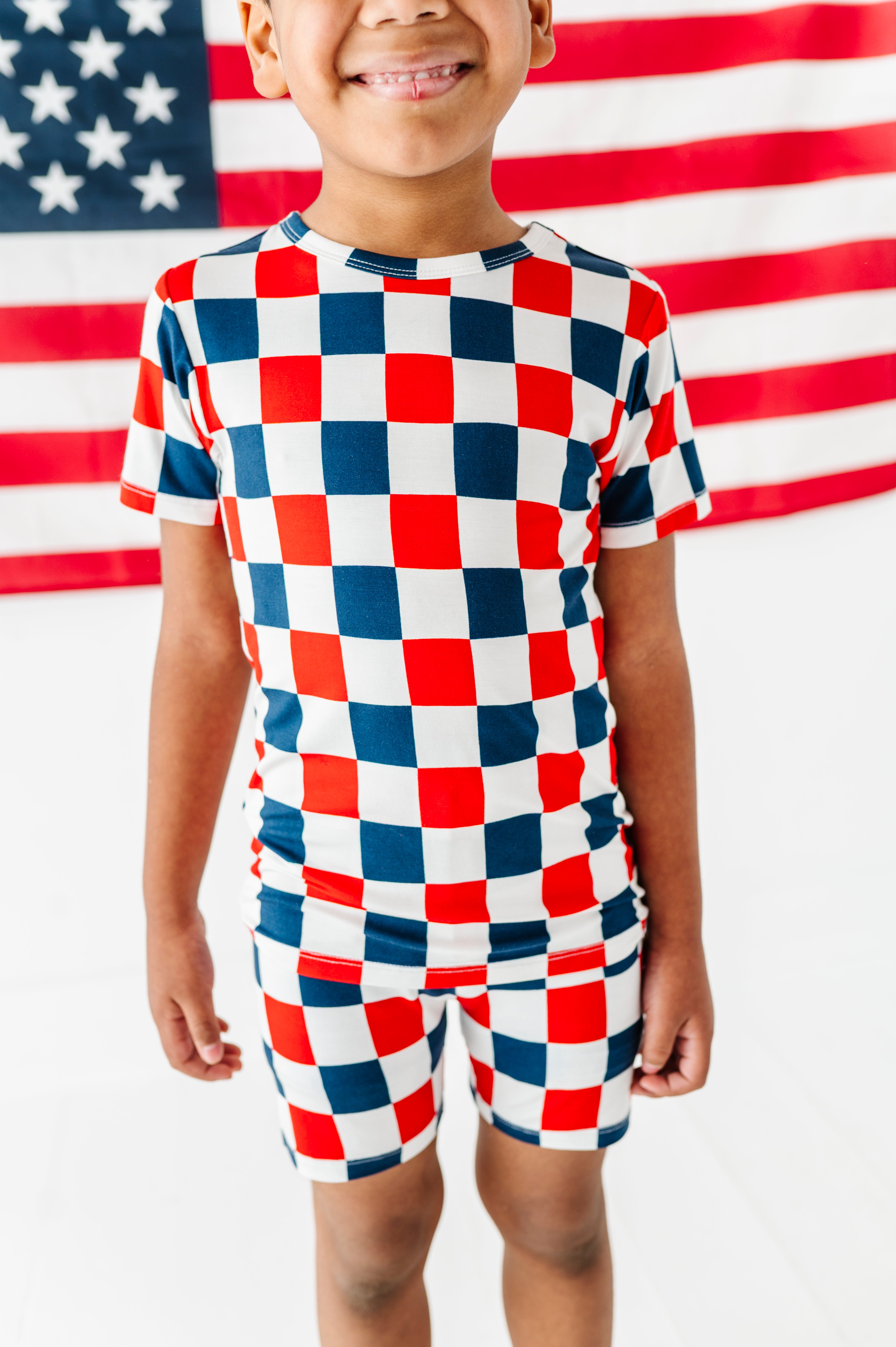 Child wearing a red, white, and blue checkered bamboo two piece pajama outfit in front of an American flag.